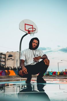 Stylish young man in urban setting squatting on a basketball court at dusk with vibrant city backdrop.