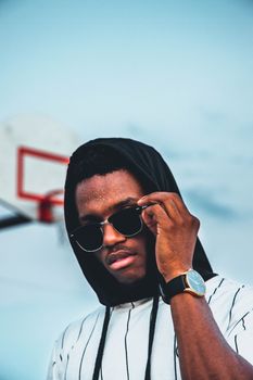 Stylish young man wearing sunglasses and a hoodie, posing by a basketball hoop outdoors.