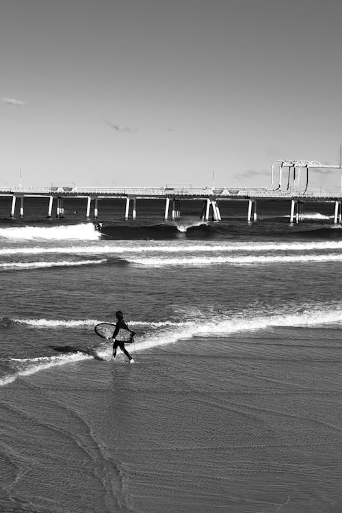 Grayscale Photo of Person Walking on Seashore Carrying a Surfboard
