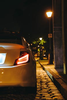 A close-up of a car's tail light on a cobblestone street, illuminated by streetlights at night.