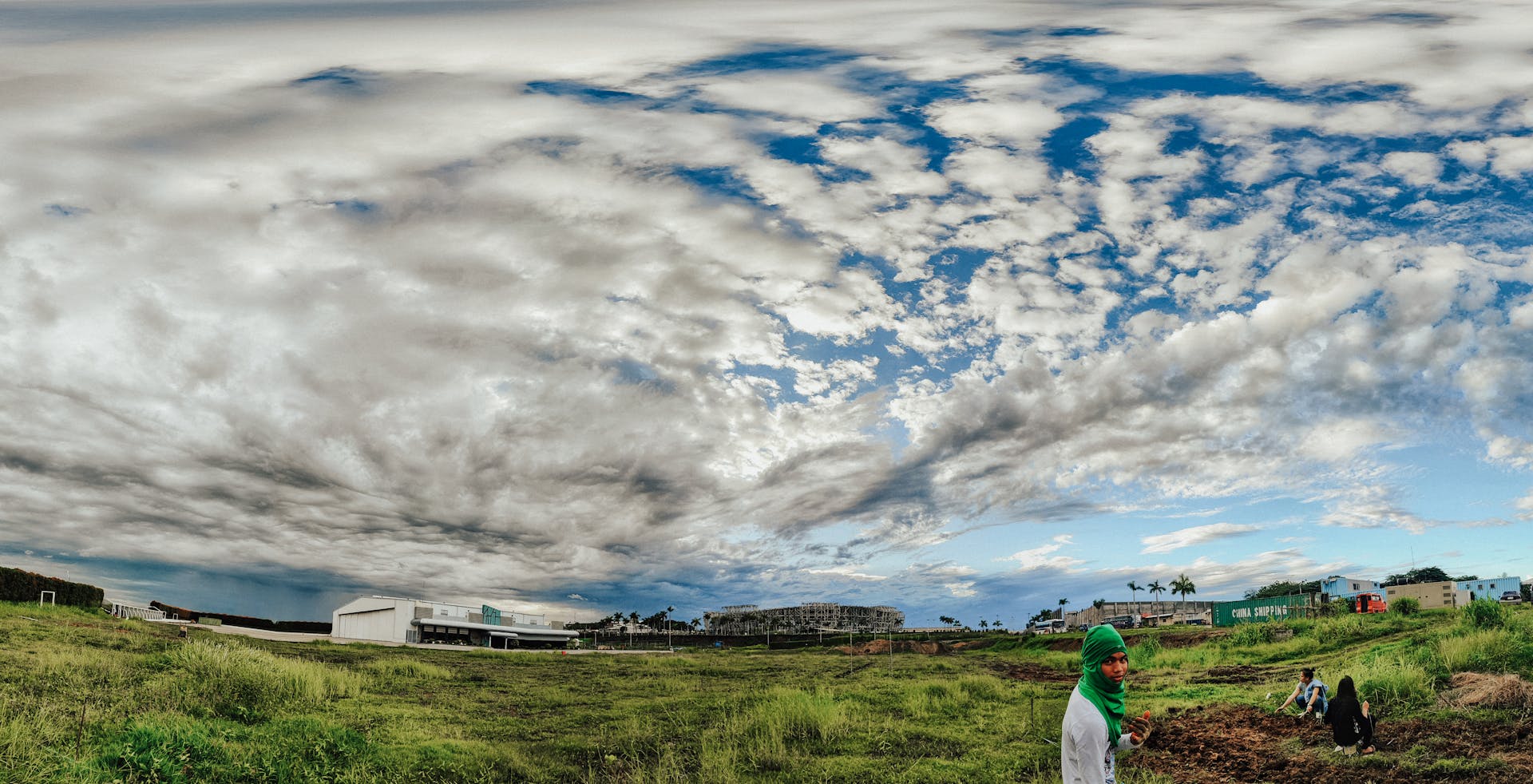 Wide-angle shot of a rural landscape featuring vibrant clouds and a vast field, with people working.