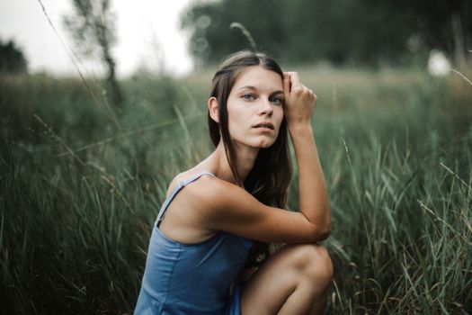 Pensive lady in dress sitting on grassy lawn with tucked leg and hand near face while looking at camera in nature with trees on blurred background