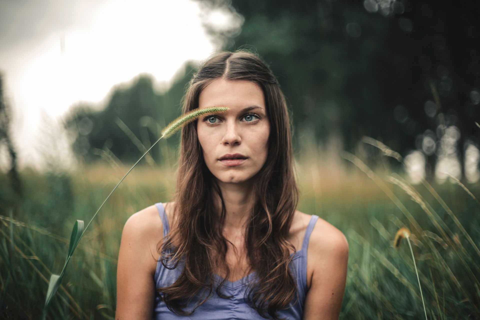Calm female with wavy hair in casual outfit looking away while sitting amidst tall grass and green trees on blurred background on summer day