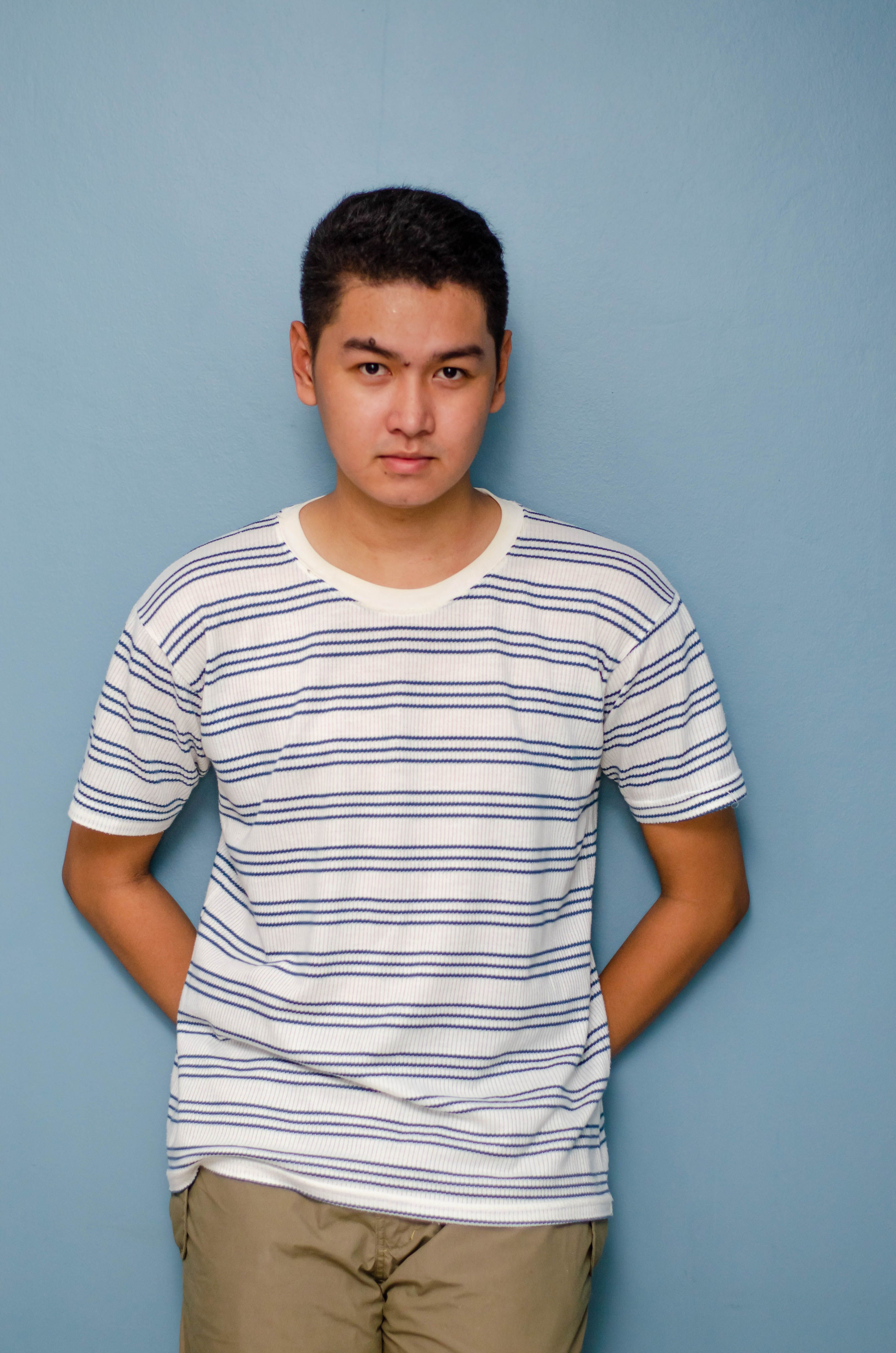 A young man in a striped shirt confidently poses against a solid blue background in studio lighting.