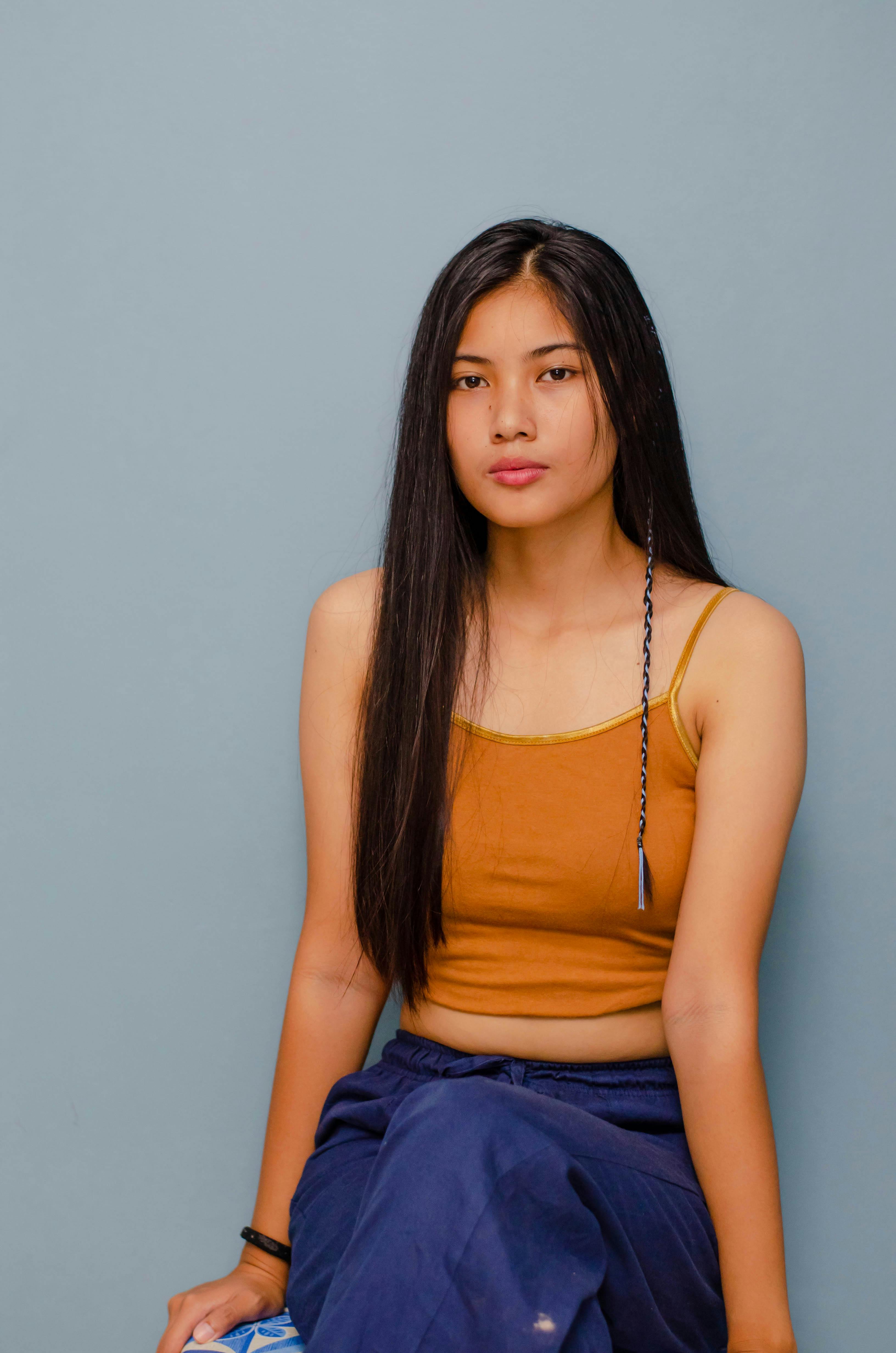Portrait of a young woman with long hair, wearing a tank top, sitting against a blue background.