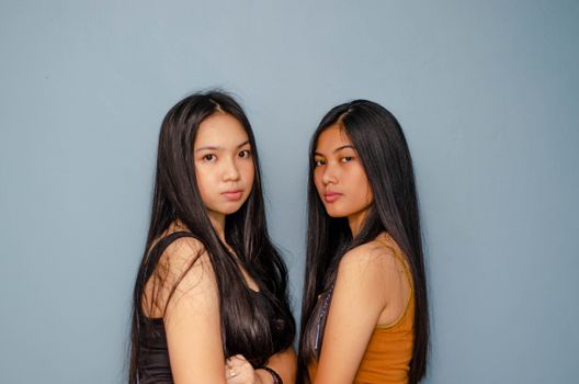 A stylish portrait of two young women with long black hair posing against a blue studio background.