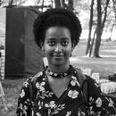 Black and white of content African American female with afro hairstyle in casual clothes looking at camera while standing in nature with trees and tent on background