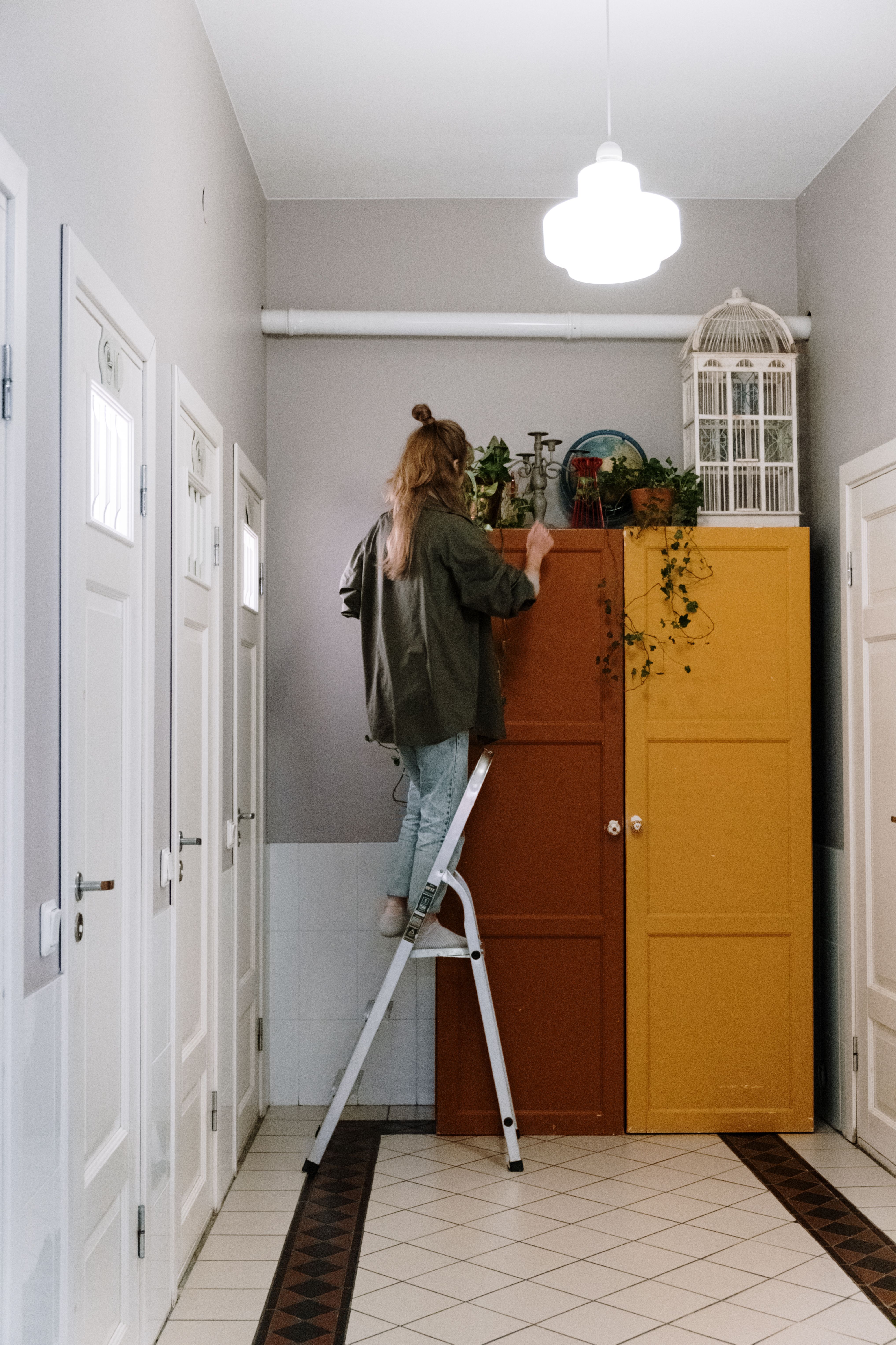 Woman standing on a silver step ladder in a bright hallway, reaching the top of a mustard and brown wardrobe