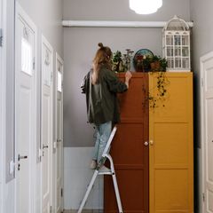 Woman standing on a silver step ladder in a bright hallway, reaching the top of a mustard and brown wardrobe