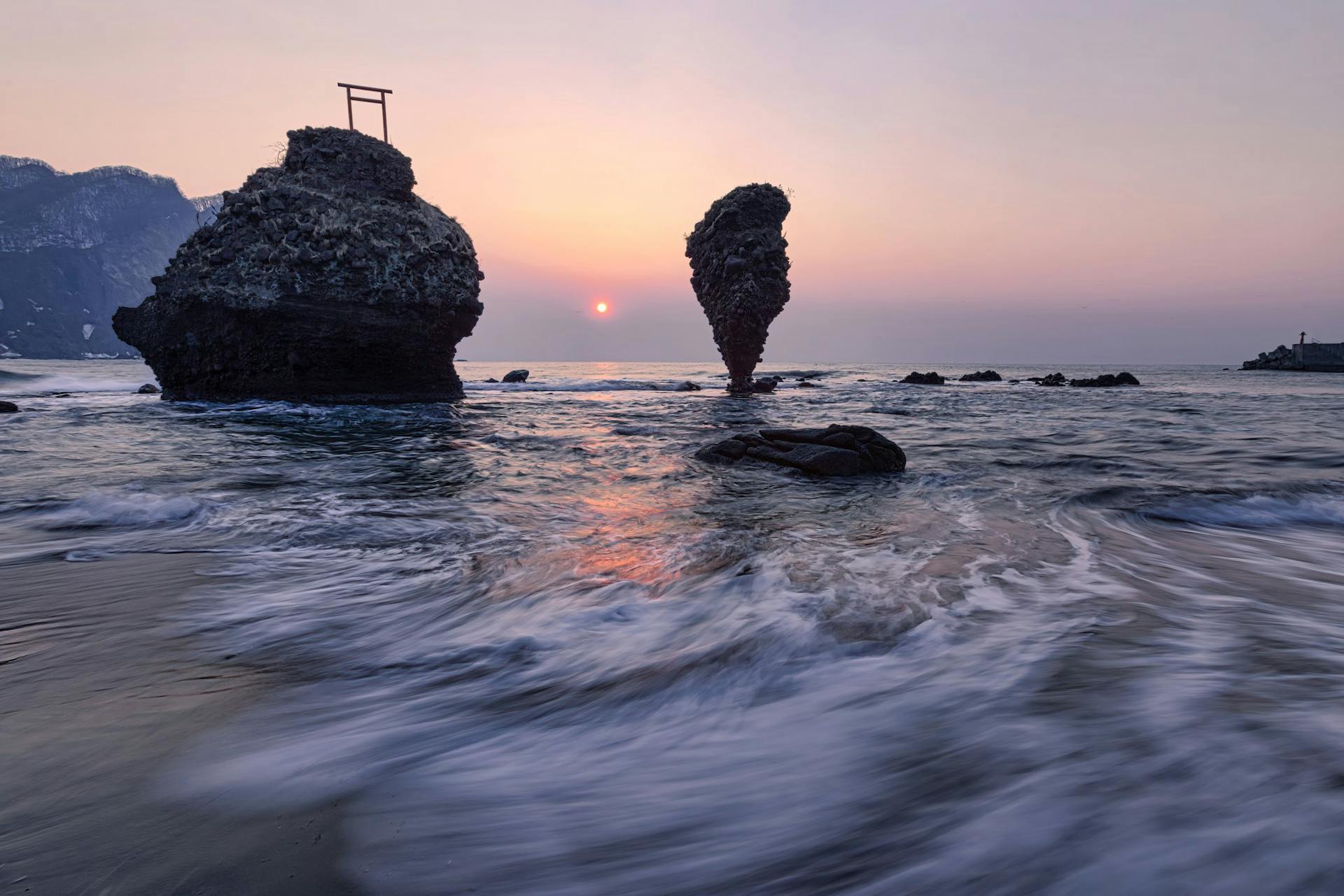 Long exposure of small vibrant Sun in light blue pink sky over mountain range and cliffs next to coast of endless wavy ocean in evening at dawn