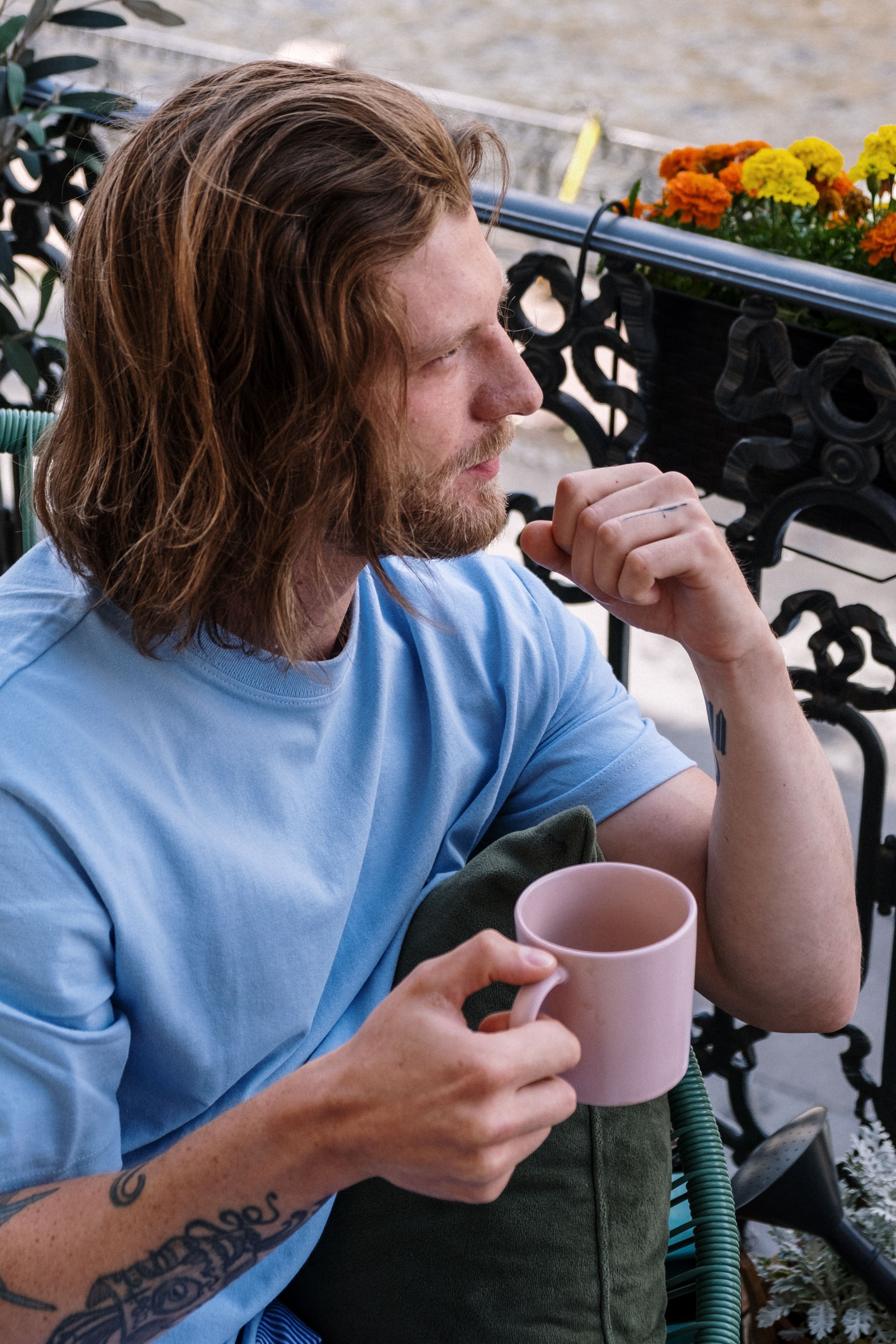 一位留着长发的男子在阳台上悠闲地喝着咖啡，若有所思，周围的花朵生机盎然