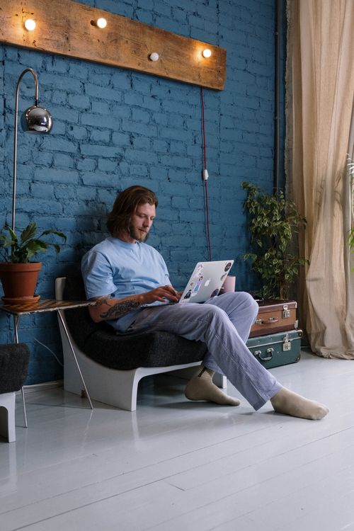 Free A bearded man with long hair using a laptop while sitting indoors in a modern, cozy living room. Stock Photo