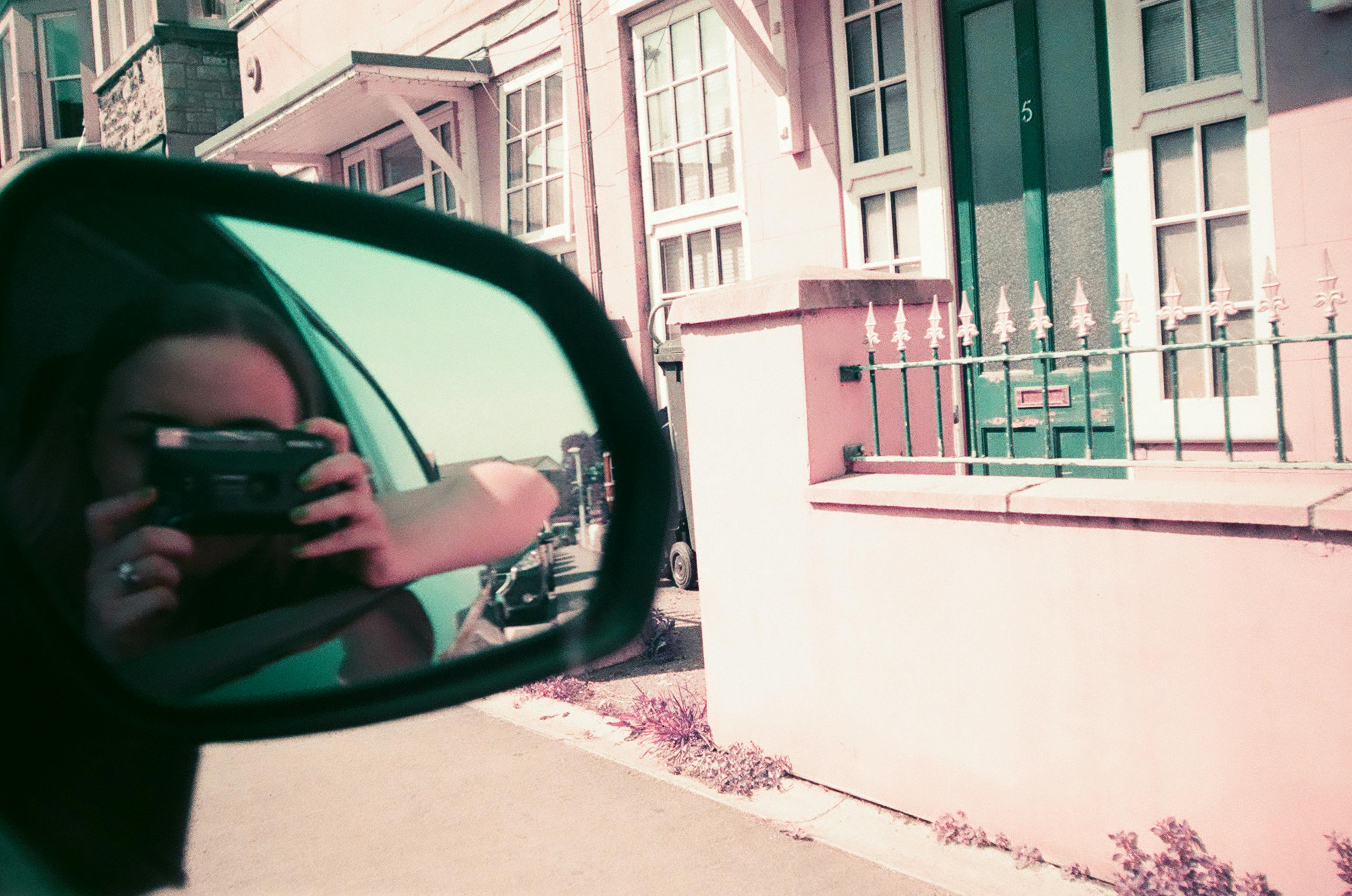 A person taking a picture of themselves using a side mirror reflection outdoors.