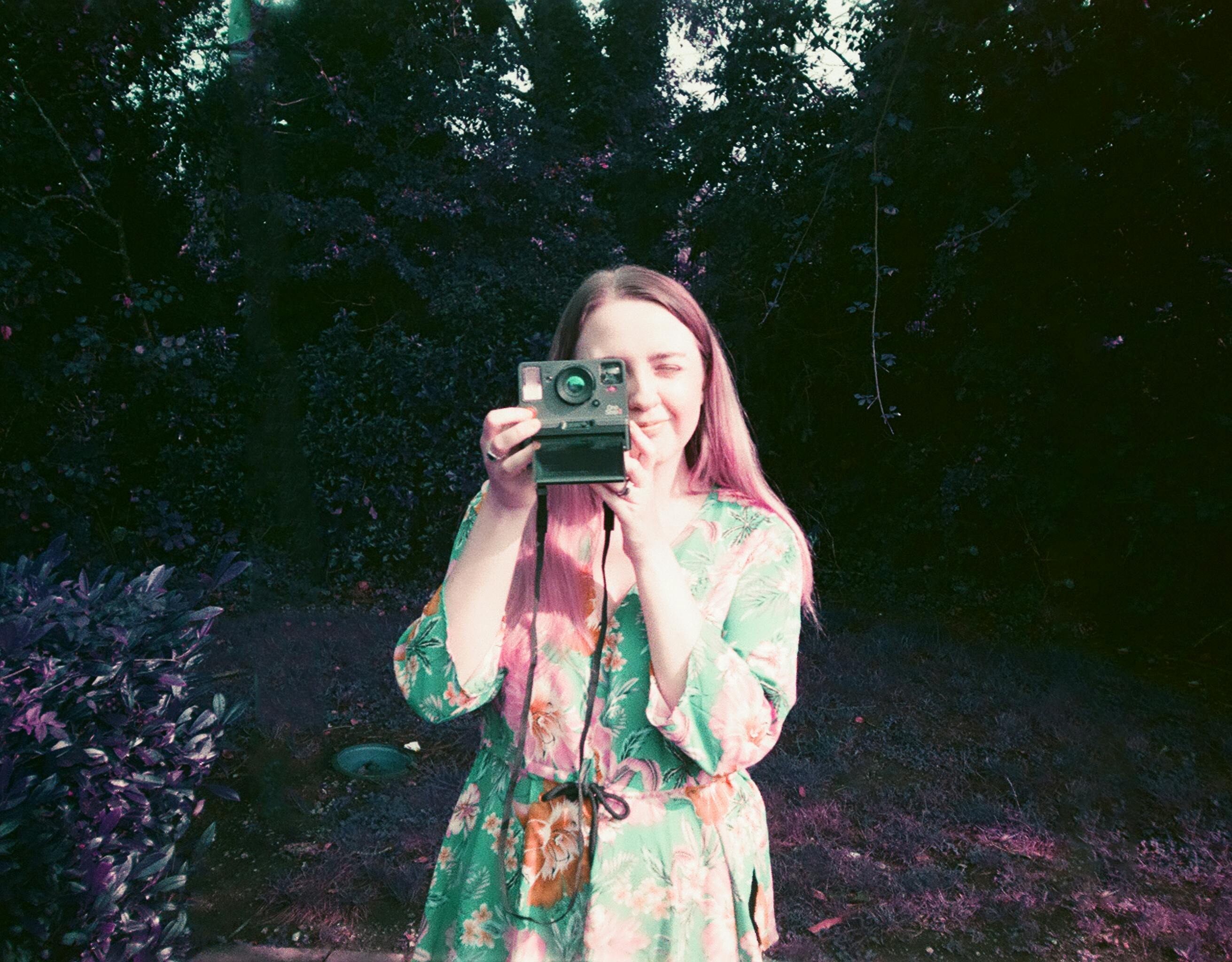 Young woman in floral dress taking a photo with a vintage camera outdoors.