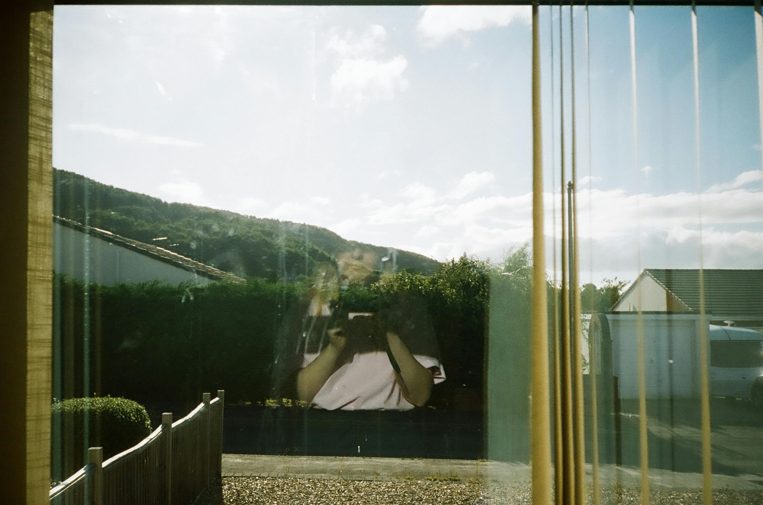 A woman takes a picture through a sunny window reflecting hills and houses.
