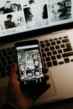 A hand holds a smartphone showing photo gallery, with a laptop screen in the background during nighttime.