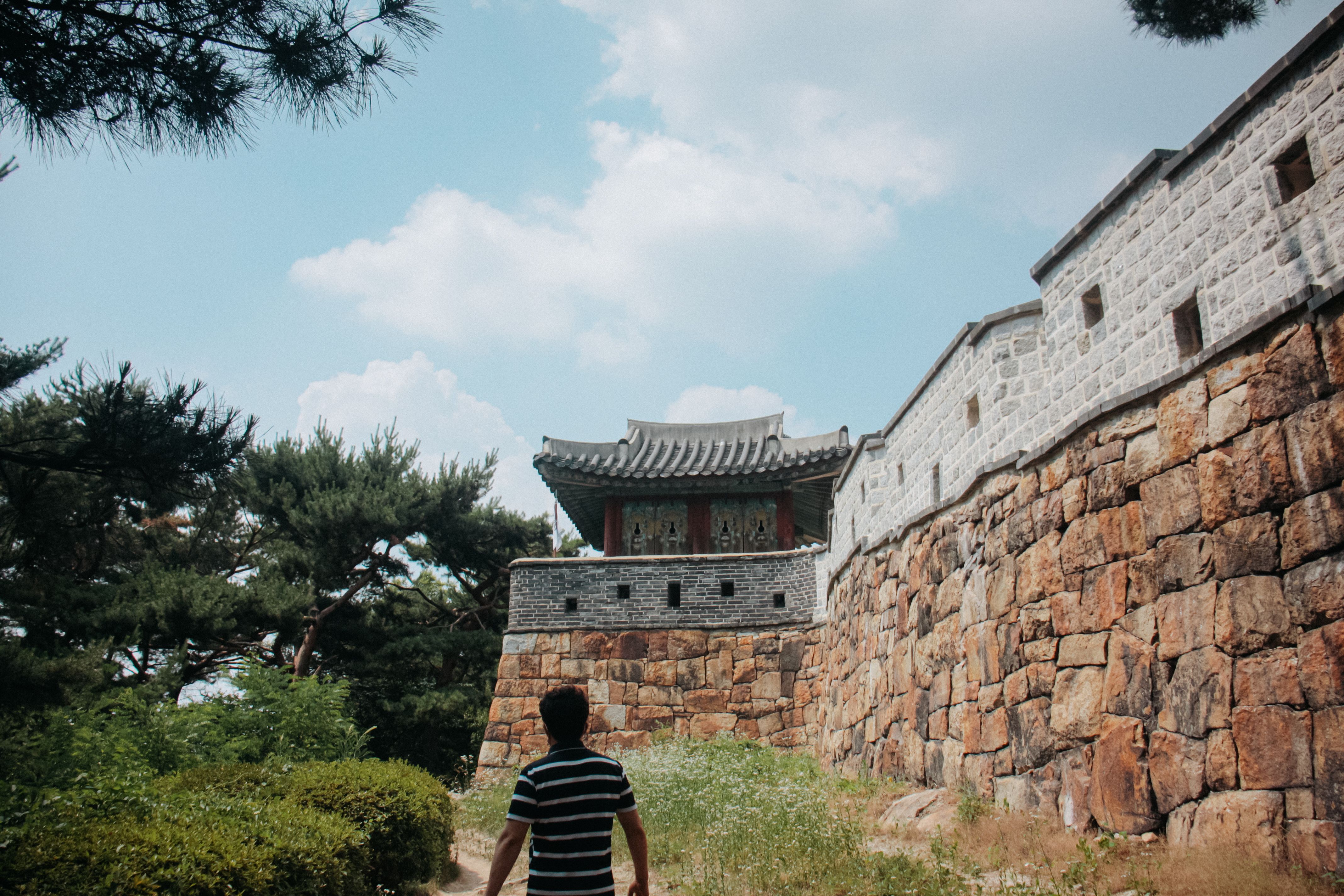A man walks towards a traditional stone fortress under a clear blue sky, surrounded by lush greenery.