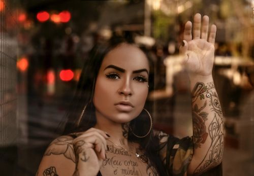 Free Through glass of attractive female with tattoos and piercing standing behind window and touching glass while looking at camera Stock Photo