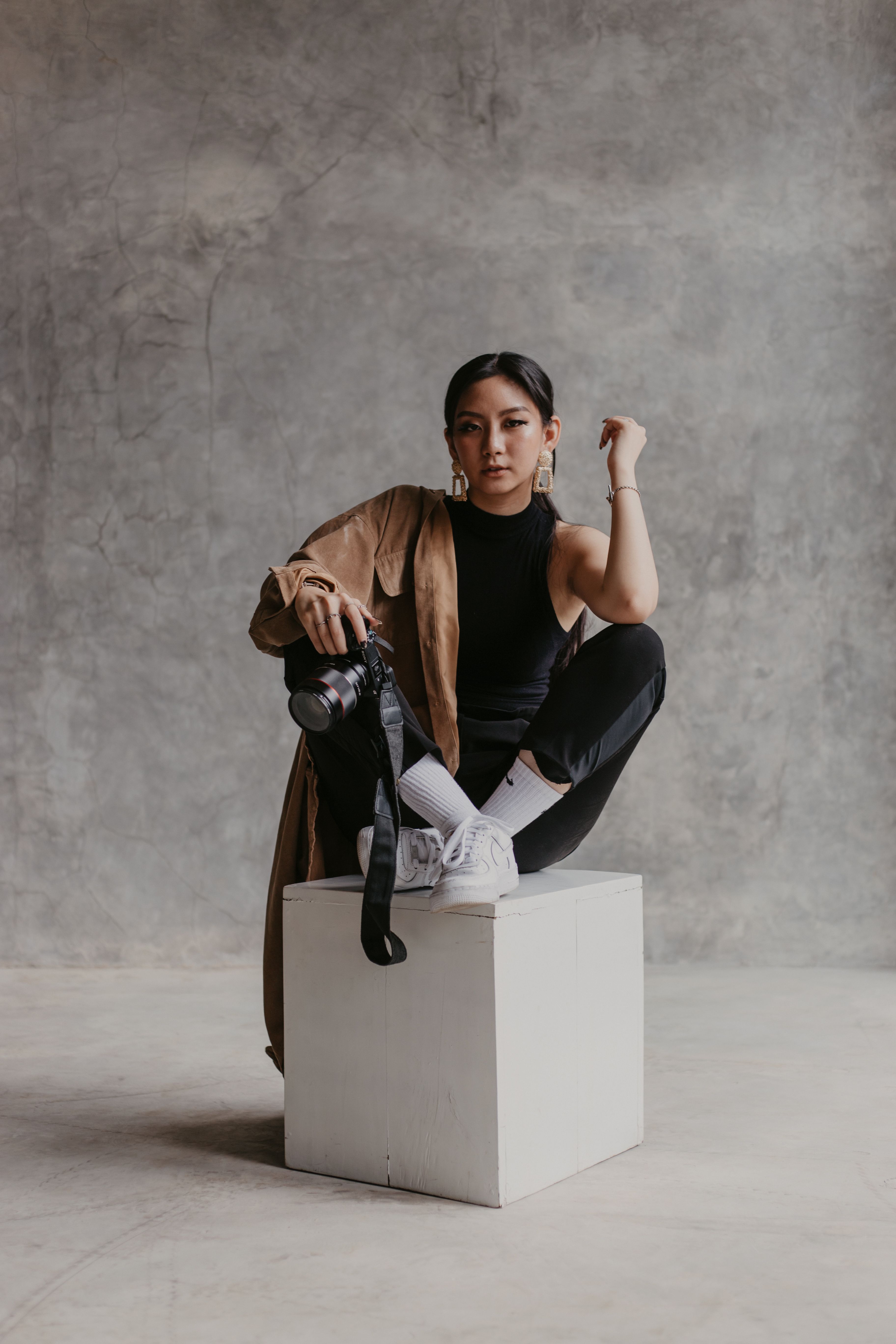 Asian woman sitting on a block in studio, holding camera, brown jacket fashion.