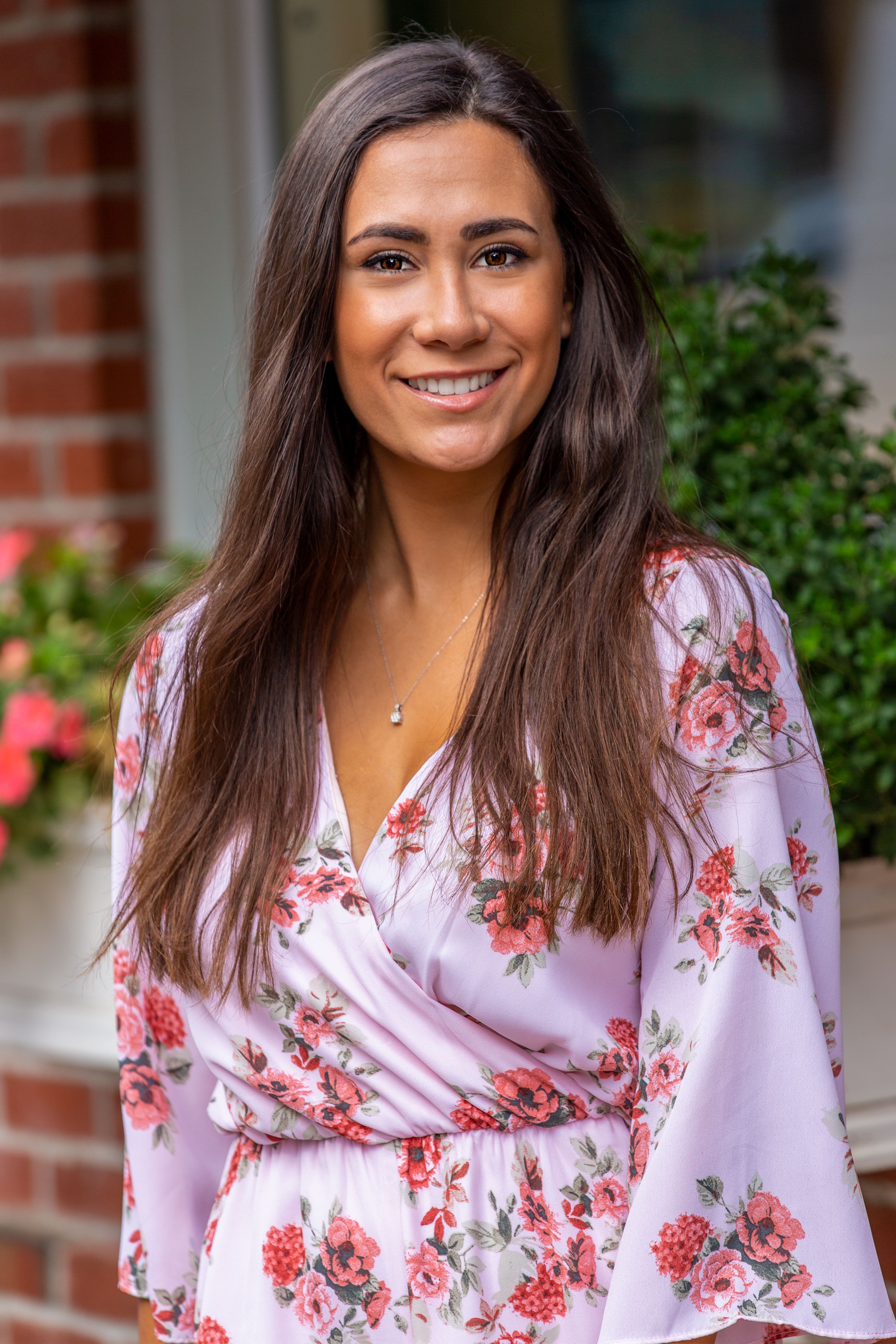Young female with long brown hair in pink dress with flowers standing outside and toothy smiling looking at camera