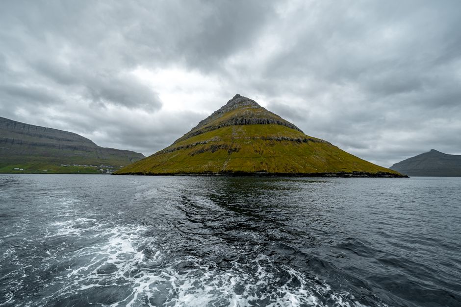Scenic view of a unique pyramid-shaped mountain rising from the ocean under a cloudy sky in the Faroe Islands.