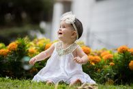 Little Baby Girl Sitting on Grass in a White Dress and Smiling 