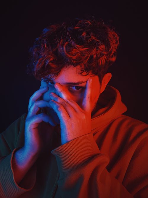 Free Insecure teen boy with curly hair wearing hoodie hiding face while standing in dark studio Stock Photo
