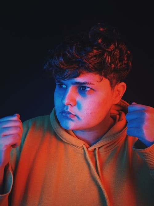 Pensive young man with curly hair and frustrated expression on face in glowing neon light  against black background