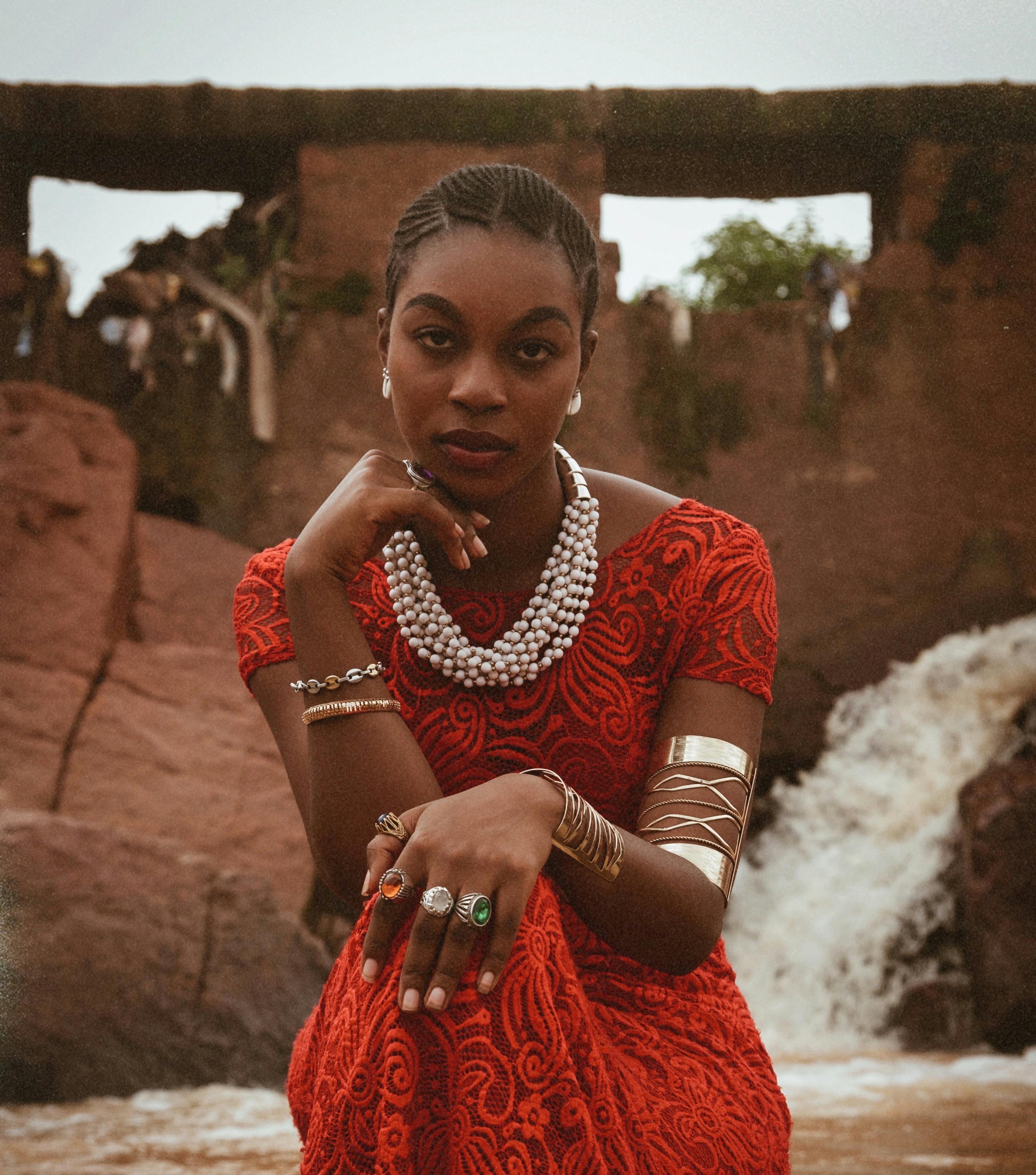 Stylish woman in a red dress poses gracefully by a waterfall, showcasing jewelry and natural beauty.