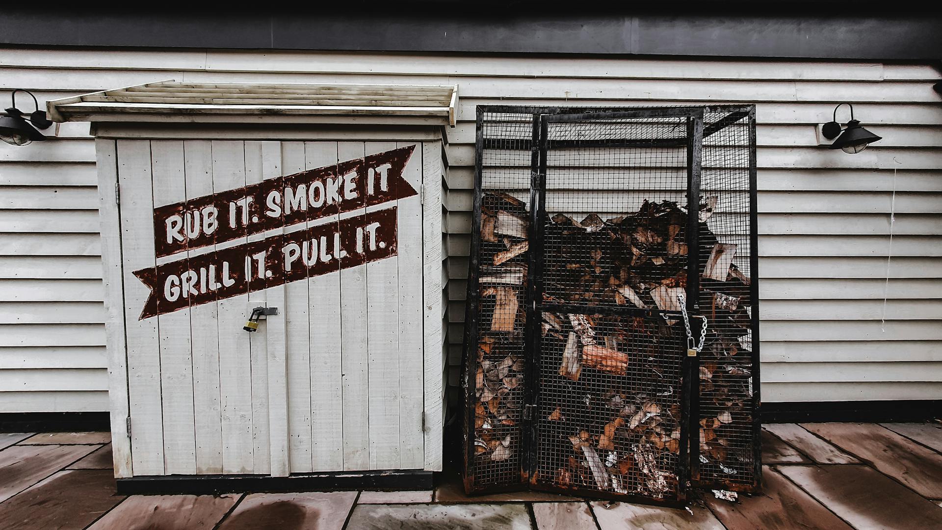 Exterior view of a rustic white shed with text alongside a metal wood storage cage.