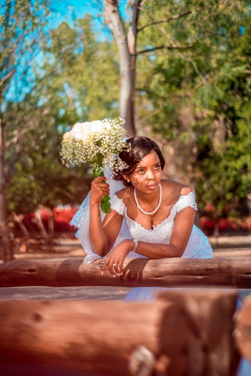 Pensive young African American bride in elegant wedding dress with bouquet of white flowers thinking on question