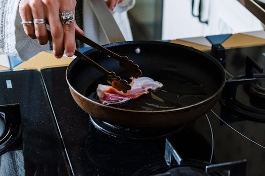 Close-up of a person cooking pork bacon with tongs in a pan on a gas stove.