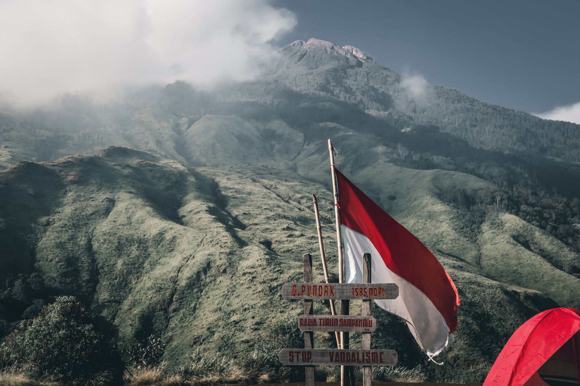 Bendera Indonesia berkibar di dekat tiang jalan dengan latar belakang gunung yang subur, melambangkan petualangan.