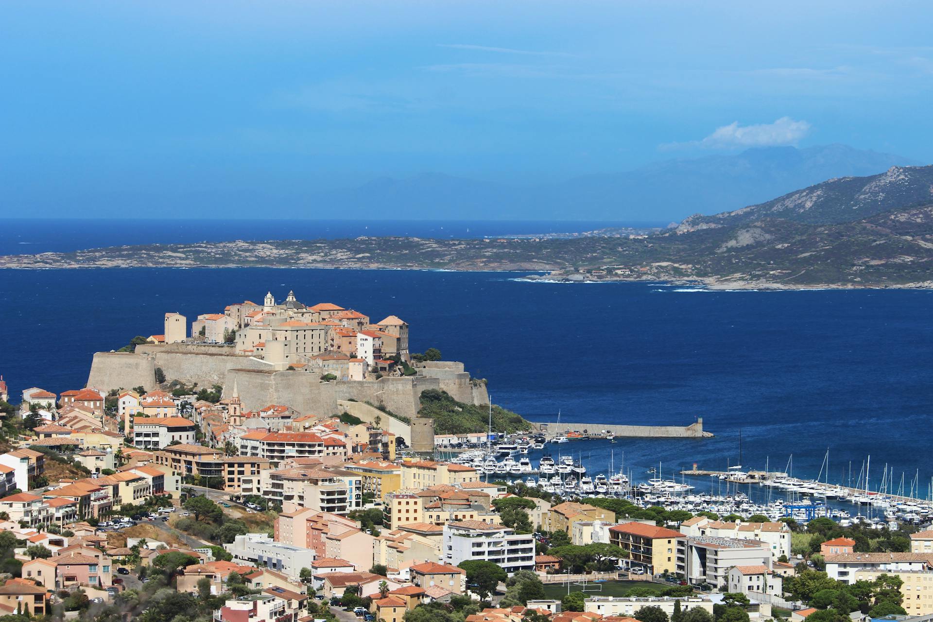 Scenic aerial view of Calvi Citadel and marina on a sunny day in Corsica, surrounded by deep blue sea and colorful houses.