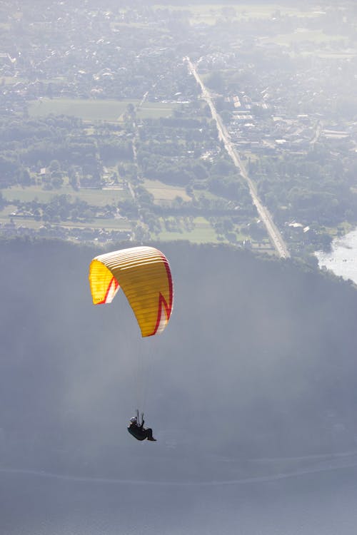 Free From above of unrecognizable person flying paraglider with yellow parachute above sea and mountain near green field with settlement in foggy weather Stock Photo