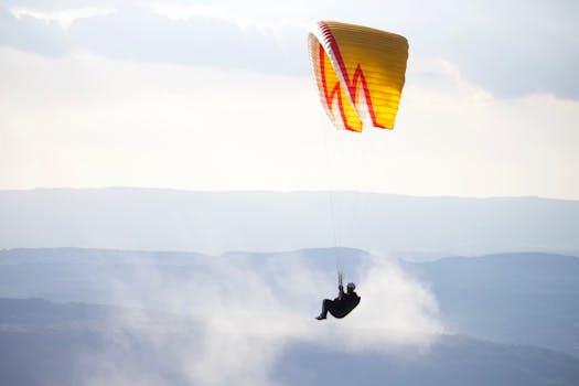 Unrecognizable person flying paraglider with yellow parachute over mountainous terrain against cloudy sky in nature in misty weather during training