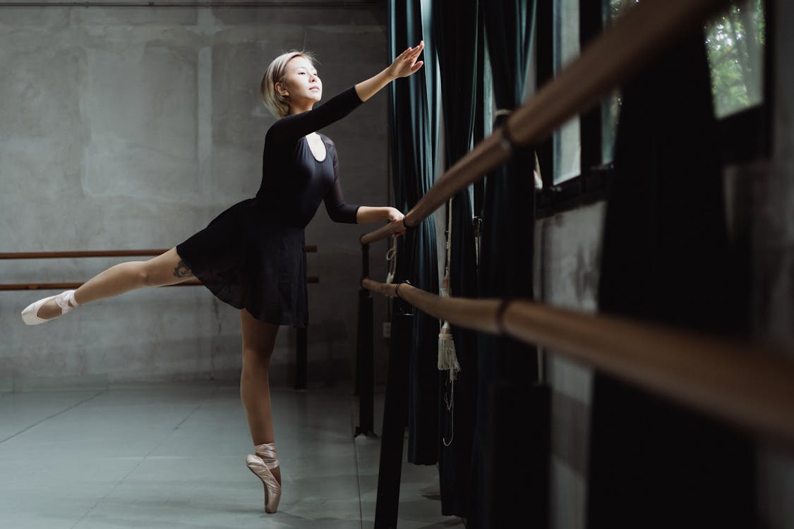 Free Full body pretty slender ballerina balancing on one leg tiptoe and raising arm gracefully near barre in modern ballet school Stock Photo