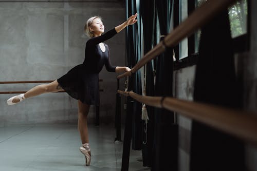 Full body pretty slender ballerina balancing on one leg tiptoe and raising arm gracefully near barre in modern ballet school