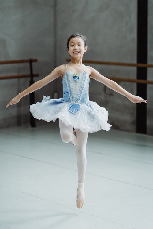 Full body of little cheerful girl with toothy smile in tutu performing ballet dancing in studio
