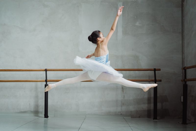 Full body back view of small ballet dancer performing split jump in studio while practicing dance