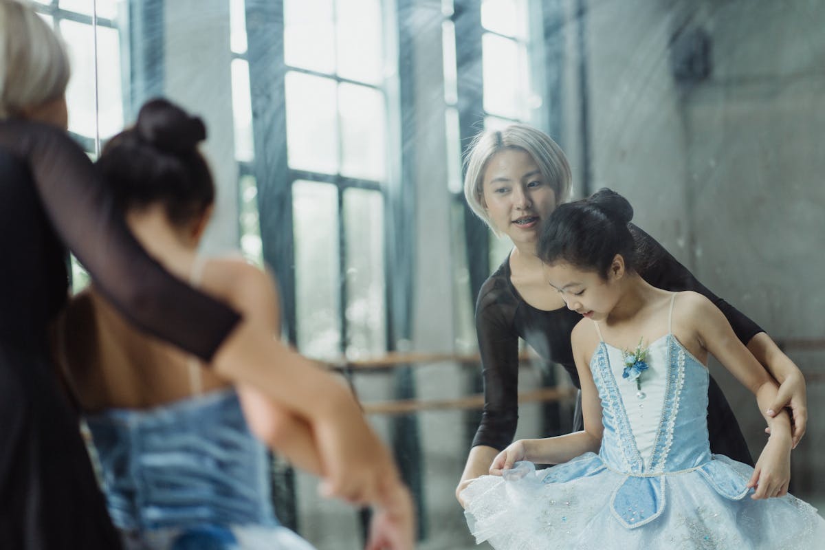 Professional ethnic ballerina teaching pupil ballet in dance room of school while looking at reflection in mirror
