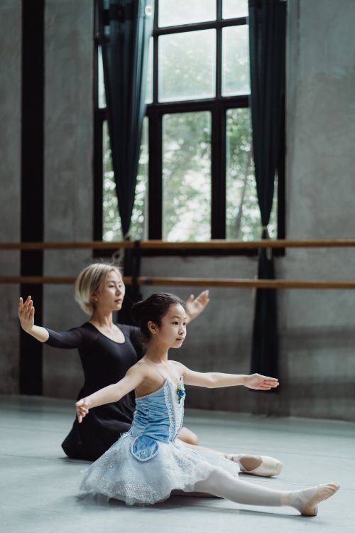 Free Full body side of ethnic little girl wearing tutu stretching legs with arms outstretched on floor with female instructor in ballet studio Stock Photo