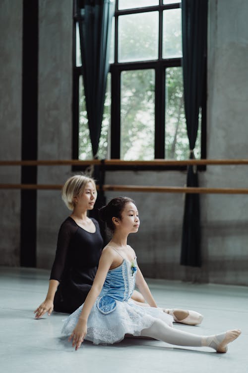 Full body of Asian girl in tutu wearing pointe shoes while sitting on floor with teacher during stretches in ballet studio