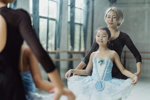 Free Female teacher standing behind back of Asian student in tutu while looking at reflection in big mirror in ballet studio Stock Photo