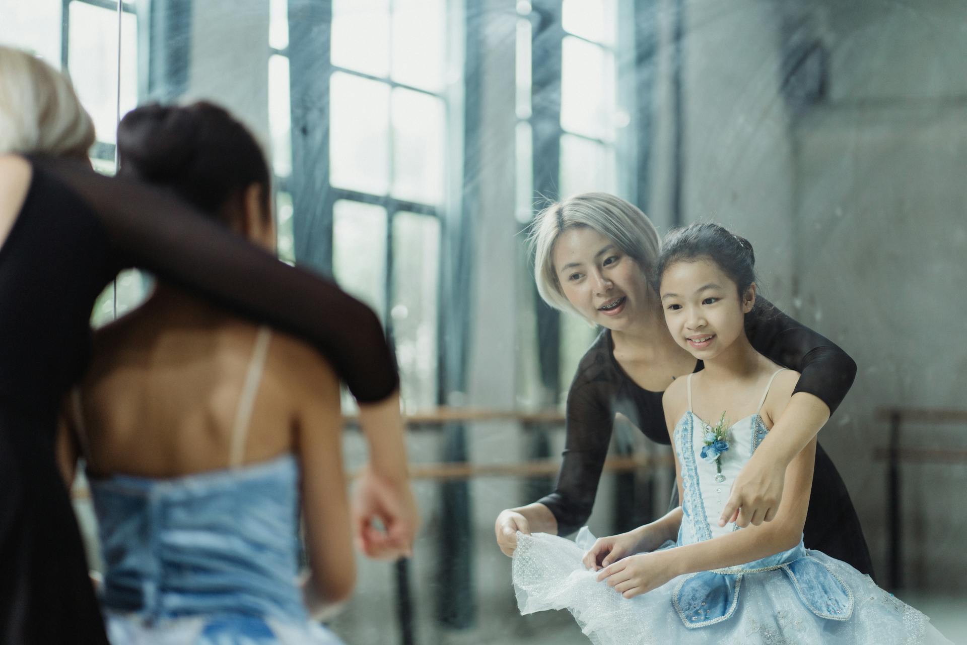 Cheerful Asian female teacher adjusting tutu of student while looking at reflection of big mirror in ballet room with barre