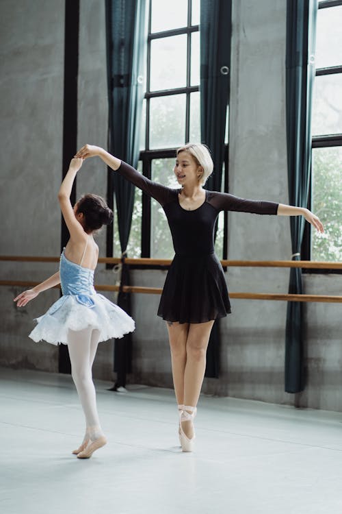 Full body of Asian teacher in black outfit and pointe shoes holding hand of female student while practicing dance movement