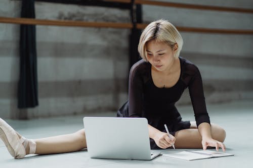 Full body of focused Asian ballerina wearing pointe shoes sitting on floor in studio with tucked leg and taking notes in notebook while looking at netbook and preparing for class