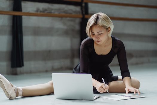 Full body of focused Asian ballerina wearing pointe shoes sitting on floor in studio with tucked leg and taking notes in notebook while looking at netbook and preparing for class