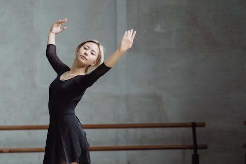 Graceful female ballet dancer wearing black outfit performing dance while standing with raised arms in studio with barre against wall