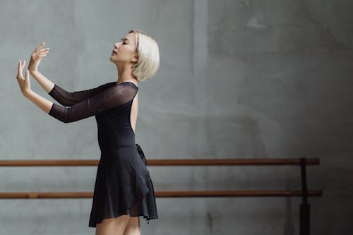 Side view of fit female ballet dancer wearing black tutu and performing dance movement with outstretched arms in ballet class